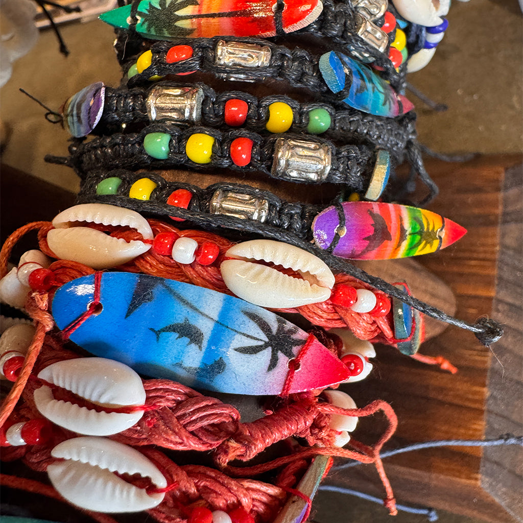 Assorted Indonesian bracelets with colorful surfboard charms, cowrie shells, and glass beads on woven cords.